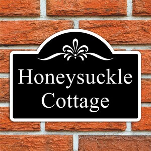 May include: A black sign with white text reading "Honeysuckle Cottage", set against a red brick wall. The sign has a decorative top with a floral design and a white border.