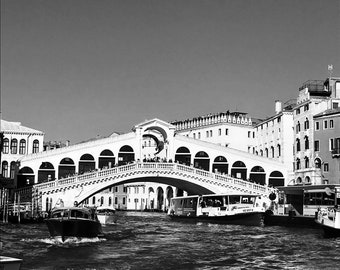 May include: A black and white photo of a bridge over a canal in Venice, Italy. The bridge is arched and has many windows. There are boats in the canal, including a gondola.