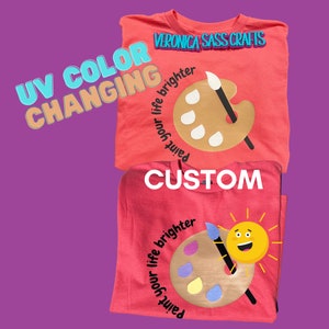 May include: Two pink t-shirts with a paint palette and paintbrush design. The text on the shirts reads "Paint your life brighter" and "Veronica Sass Crafts". The shirts are made with UV color changing fabric.