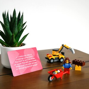 Może przedstawiać: R&oacute;żowa karta z tekstem "Stw&oacute;rz sw&oacute;j pomysł lub wyzwanie, używając LEGO, Play-Doh lub rysunk&oacute;w wykonanych na papierze. Baw się swoim modelem. Co możesz dodać, usunąć lub zmienić, aby ulepszyć rzeczy?" Mały ż&oacute;łty koparkowy pojazd zabawka, mała czerwona taczka zabawka i mała brązowo-ż&oacute;łta konstrukcja z klock&oacute;w zabawka znajdują się na drewnianej powierzchni. Mała zielona roślina sukulentna znajduje się w białym doniczce.