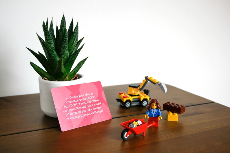 Może przedstawiać: R&oacute;żowa karta z tekstem "Stw&oacute;rz sw&oacute;j pomysł lub wyzwanie, używając LEGO, Play-Doh lub rysunk&oacute;w wykonanych na papierze. Baw się swoim modelem. Co możesz dodać, usunąć lub zmienić, aby ulepszyć rzeczy?" Mały ż&oacute;łty koparkowy pojazd zabawka, mała czerwona taczka zabawka i mała brązowo-ż&oacute;łta konstrukcja z klock&oacute;w zabawka znajdują się na drewnianej powierzchni. Mała zielona roślina sukulentna znajduje się w białym doniczce.