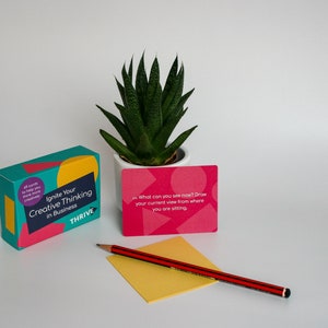 Może przedstawiać: Pudełko kart z tekstem "Ignite Your Creative Thinking in Business" i słowem "THRIVE" na przodzie. Jedna karta została wyciągnięta z pudełka i zawiera napis "What can you see now? Draw your current view from where you are sitting". Na zdjęciu widoczny jest r&oacute;wnież oł&oacute;wek i ż&oacute;łty notes.