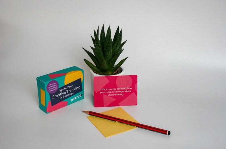 Może przedstawiać: Pudełko kart z tekstem "Ignite Your Creative Thinking in Business" i słowem "THRIVE" na przodzie. Jedna karta została wyciągnięta z pudełka i zawiera napis "What can you see now? Draw your current view from where you are sitting". Na zdjęciu widoczny jest r&oacute;wnież oł&oacute;wek i ż&oacute;łty notes.