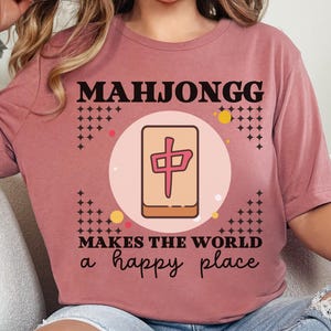 Könnte beinhalten: Mauvefarbenes T-Shirt mit dem Wort "MAHJONGG" und einer Mahjong-Stein-Grafik. Der Text "MAKES THE WORLD a happy place" ist darunter gedruckt. Das T-Shirt ist im lässigen Stil gehalten und für den Alltag geeignet.