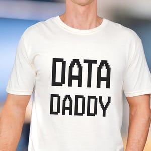 May include: A white t-shirt with the text "DATA DADDY" in a pixelated black font. The shirt is short-sleeved and appears to be made of a soft material. The person is wearing light-wash jeans.
