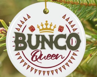 Puede incluir: Adorno navideño redondo y blanco con la inscripción "BUNCO Queen" en verde oscuro y burdeos. Presenta una corona dorada, dados y detalles triangulares. Una cuerda dorada está unida para colgarlo.