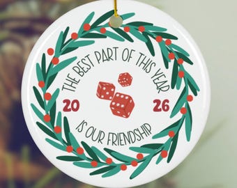 May include: A round white ceramic ornament featuring a green wreath with red berries. In the center, three red dice are illustrated with the text 'THE BEST PART OF THIS YEAR IS OUR FRIENDSHIP' and the numbers '20' and '26'. It hangs by a gold string.