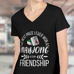 May include: Black V-neck t-shirt with the text "The best prize I ever won at Mahjong is our friendship" in white. The design includes a Mahjong tile graphic.