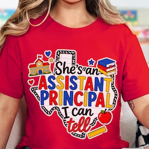 May include: A red t-shirt featuring a graphic of the state of Texas filled with lined paper texture. The text reads "She's an ASSISTANT PRINCIPAL I can tell" in blue, yellow, and red. Icons include a school building, books, an apple, and a ruler.