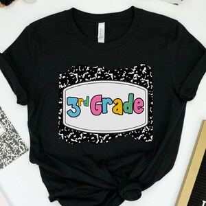 May include: Black t-shirt featuring a colorful "3rd Grade" graphic. The design is set within a white-bordered rectangle, surrounded by a black and white composition notebook-style border. The shirt is folded, showing its texture.