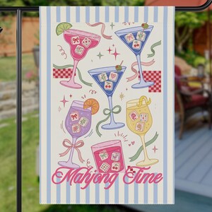 May include: A garden flag featuring a whimsical illustration of various cocktails, each adorned with mahjong tiles and garnishes. The flag has a cream background with blue vertical stripes and the words "Mahjong Time" in a decorative font.