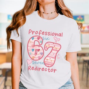 May include: White t-shirt with the text "Professional Six 67 Seven Redirector" in pink and red. The number 67 is decorated with hearts and stripes. The shirt is a crew neck style.