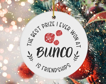 Puede incluir: Adorno navideño redondo blanco con el texto "El mejor premio que he ganado en Bunco son las amistades" y un dibujo de tres dados rojos.
