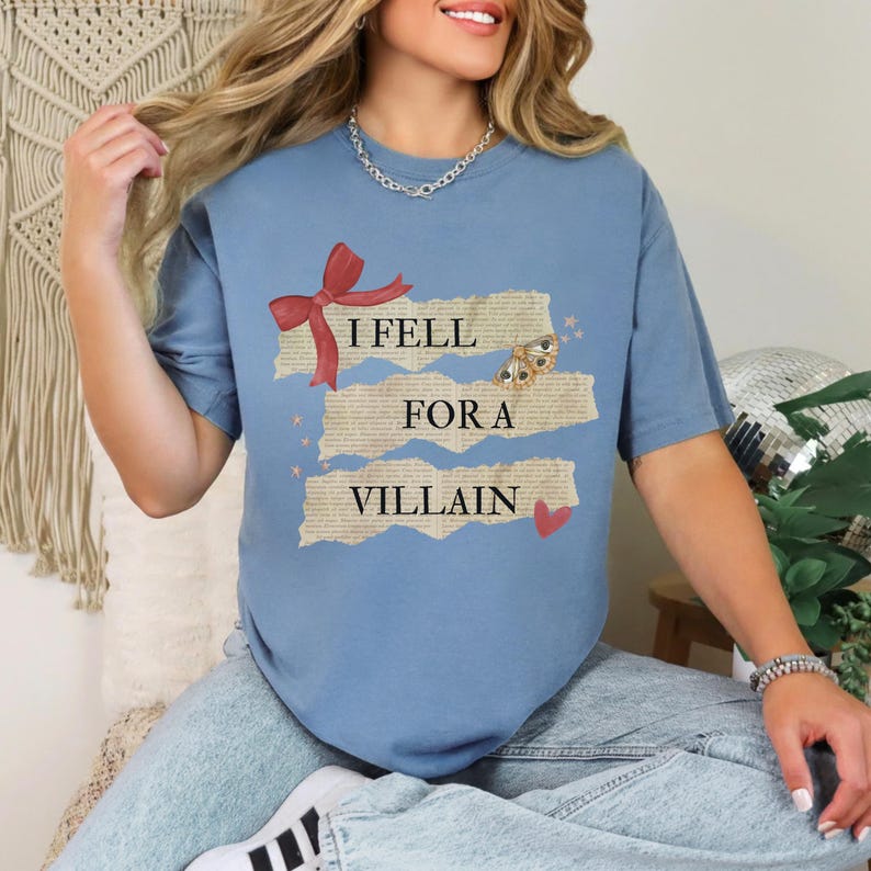 K&ouml;nnte beinhalten: Hellblaues T-Shirt mit dem Text "I Fell For A Villain". Das Design umfasst eine rote Schleife, einen Schmetterling und ein kleines rotes Herz. Das T-Shirt hat einen l&auml;ssigen Stil.