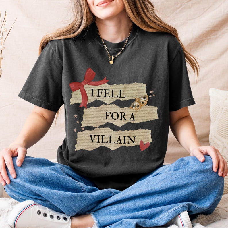 K&ouml;nnte beinhalten: Dunkelgraues T-Shirt mit dem Text "I Fell For A Villain" in einem Used-Look-Design. Die Grafik enth&auml;lt eine rote Schleife, einen Schmetterling und ein Herz. Das Shirt ist ein l&auml;ssiger Stil mit kurzen &Auml;rmeln.