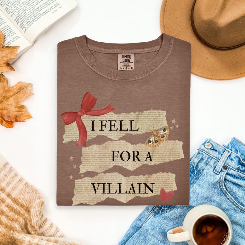 K&ouml;nnte beinhalten: Braunes T-Shirt mit dem Text "I Fell For A Villain". Das Design umfasst eine rote Schleife, einen Schmetterling und ein Herz. Das T-Shirt liegt gefaltet auf einer wei&szlig;en Oberfl&auml;che.