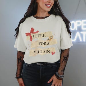 K&ouml;nnte beinhalten: Cremefarbenes T-Shirt mit dem Text "I Fell For A Villain" in einem geschichteten Design. Das Design umfasst eine rote Schleife, einen Schmetterling und ein Herz. Das Shirt hat kurze &Auml;rmel.