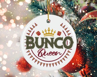 Puede incluir: Adorno navideño redondo de cerámica blanca con el texto "Bunco Queen" en marrón y burdeos. El adorno presenta una corona dorada y gráficos de dados, suspendido por un hilo dorado. Fondo borroso de un árbol de Navidad con adornos rojos.