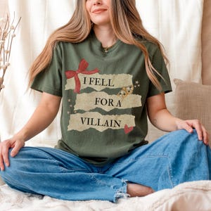 K&ouml;nnte beinhalten: Olivgr&uuml;nes T-Shirt mit dem Text "I Fell For A Villain". Das Design umfasst eine rote Schleife, einen Schmetterling, Sterne und ein Herz. Das Shirt hat kurze &Auml;rmel und einen Rundhalsausschnitt.