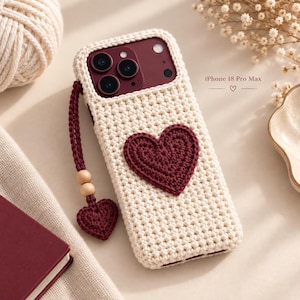 Può includere: Una custodia per telefono all'uncinetto color crema per iPhone 18 Pro Max con un'applicazione a forma di cuore rosso scuro sul retro. È fissato un cinturino da polso all'uncinetto rosso scuro abbinato, decorato con due perline di legno.