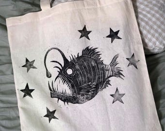 May include: Cream-colored tote bag featuring a black anglerfish design and black star accents. The bag has sturdy handles and is made of a durable fabric, perfect for carrying groceries or books.