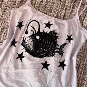 May include: A light lavender tank top featuring a black linocut-style graphic of an anglerfish surrounded by six black stars. The garment has thin spaghetti straps and a scoop neckline, showcasing a bold, high-contrast print on the front.