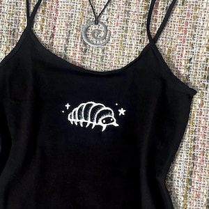 Puede incluir: Camisola negra con una ilustración blanca de un bicho bolita y dos estrellas. Un colgante de espiral plateado cuelga de un collar de cordón negro. La parte superior tiene tirantes finos.