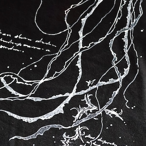 May include: A black t-shirt featuring a white graphic print of long, flowing, organic lines resembling jellyfish tentacles or seaweed. The design includes small white dots and illegible, cursive-style handwritten text integrated into the composition.