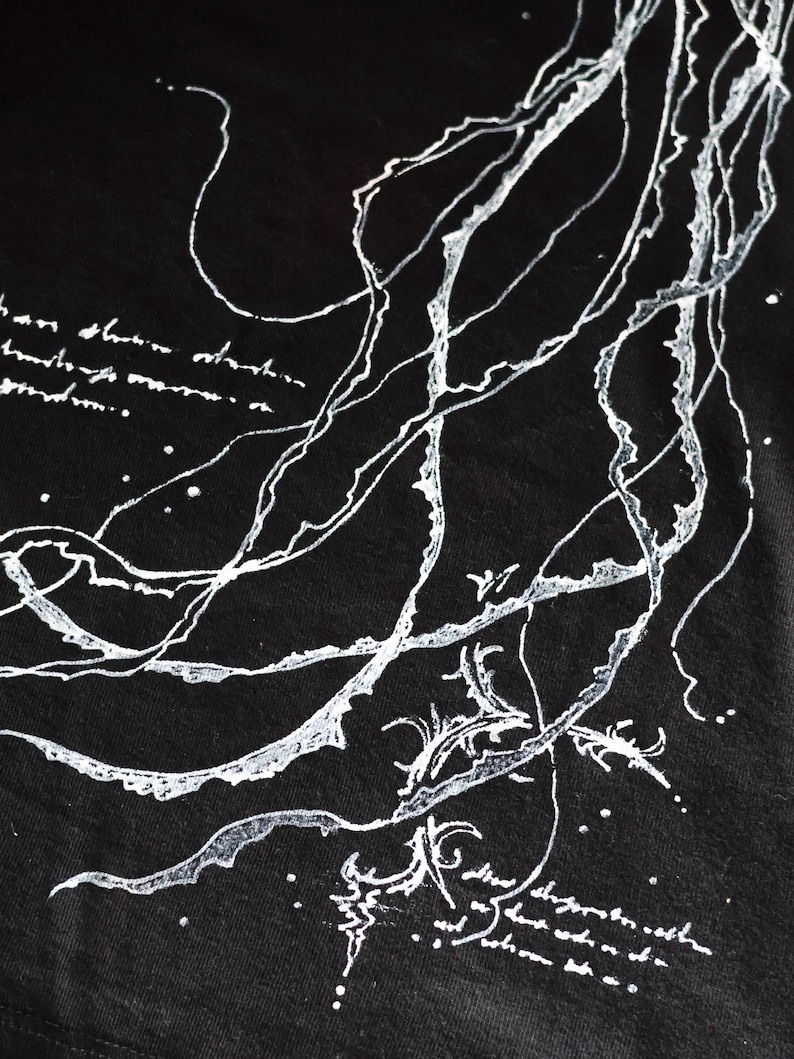 May include: A black t-shirt featuring a white graphic print of long, flowing, organic lines resembling jellyfish tentacles or seaweed. The design includes small white dots and illegible, cursive-style handwritten text integrated into the composition.