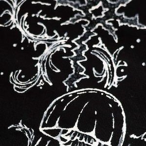 May include: A black fabric surface featuring a white graphic print of a jellyfish positioned below a large, jagged, star-like shape. The design includes intricate, swirling, vine-like patterns and small dots surrounding the central elements.
