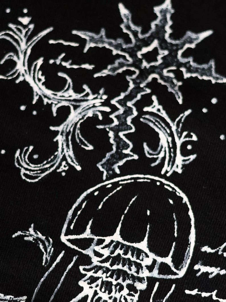 May include: A black fabric surface featuring a white graphic print of a jellyfish positioned below a large, jagged, star-like shape. The design includes intricate, swirling, vine-like patterns and small dots surrounding the central elements.