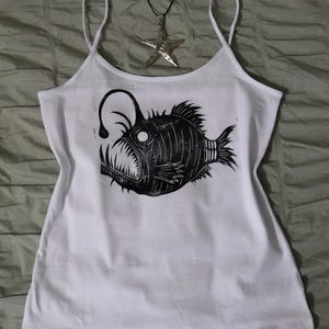 May include: A white spaghetti strap tank top featuring a black block print graphic of an anglerfish with sharp teeth and a glowing lure. A silver-toned starfish pendant on a thin black cord rests above the neckline of the tank top.