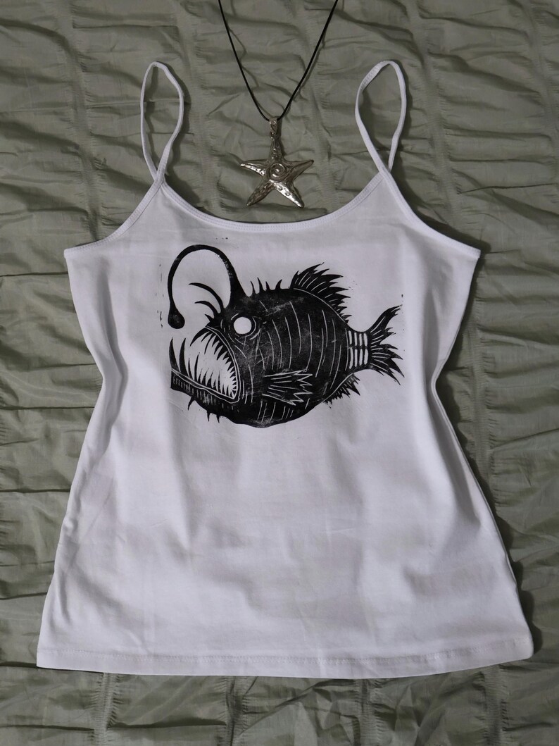 May include: A white spaghetti strap tank top featuring a black block print graphic of an anglerfish with sharp teeth and a glowing lure. A silver-toned starfish pendant on a thin black cord rests above the neckline of the tank top.