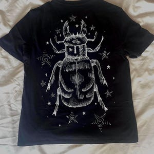 May include: Black t-shirt featuring a detailed white illustration of a beetle with large mandibles and a segmented body. The design includes stars and swirling patterns, creating a celestial theme. The shirt is made of a soft material.