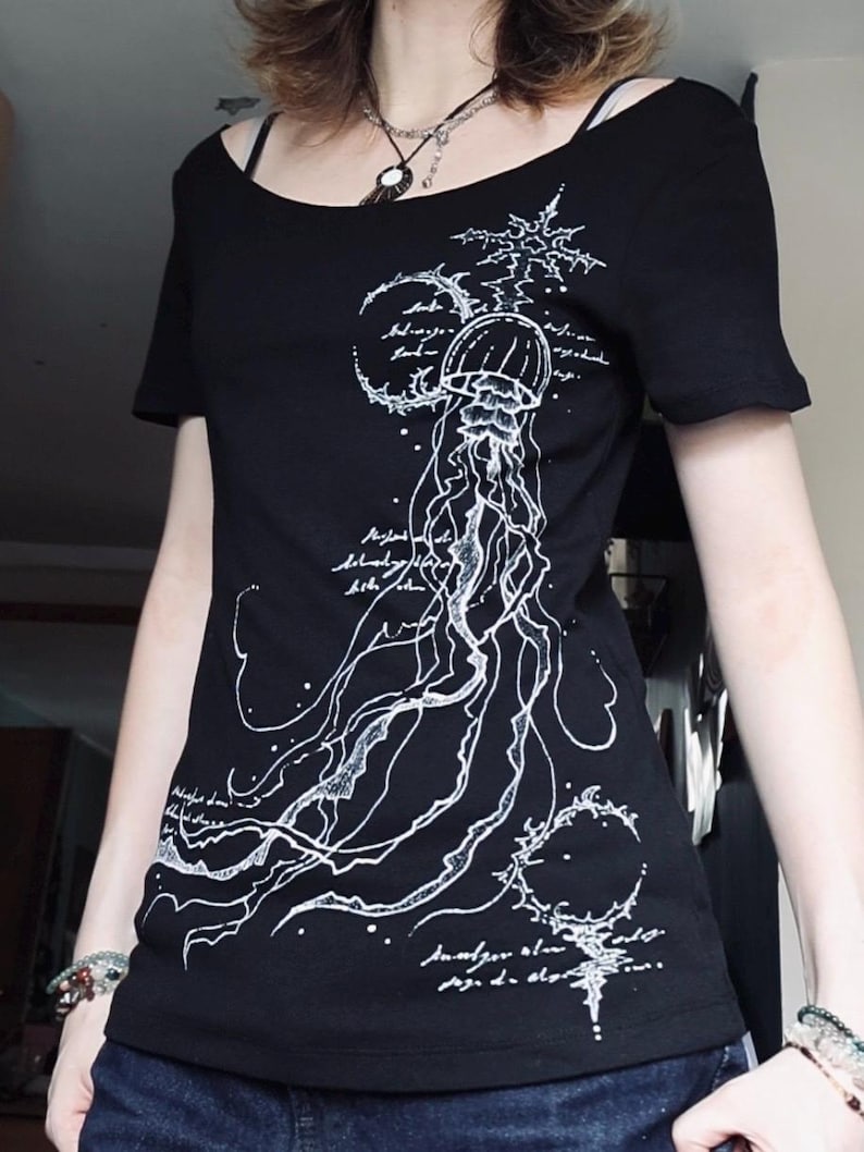 May include: A black short-sleeved t-shirt featuring a white line-art graphic of a jellyfish surrounded by celestial symbols and cursive script text. The design extends from the upper chest down to the hem, showcasing intricate tentacles and patterns.