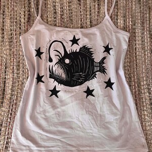 May include: A light pink tank top featuring a black graphic print of an anglerfish surrounded by six black stars. The garment has a scoop neckline and thin spaghetti straps, with the fish illustration centered on the front of the shirt.