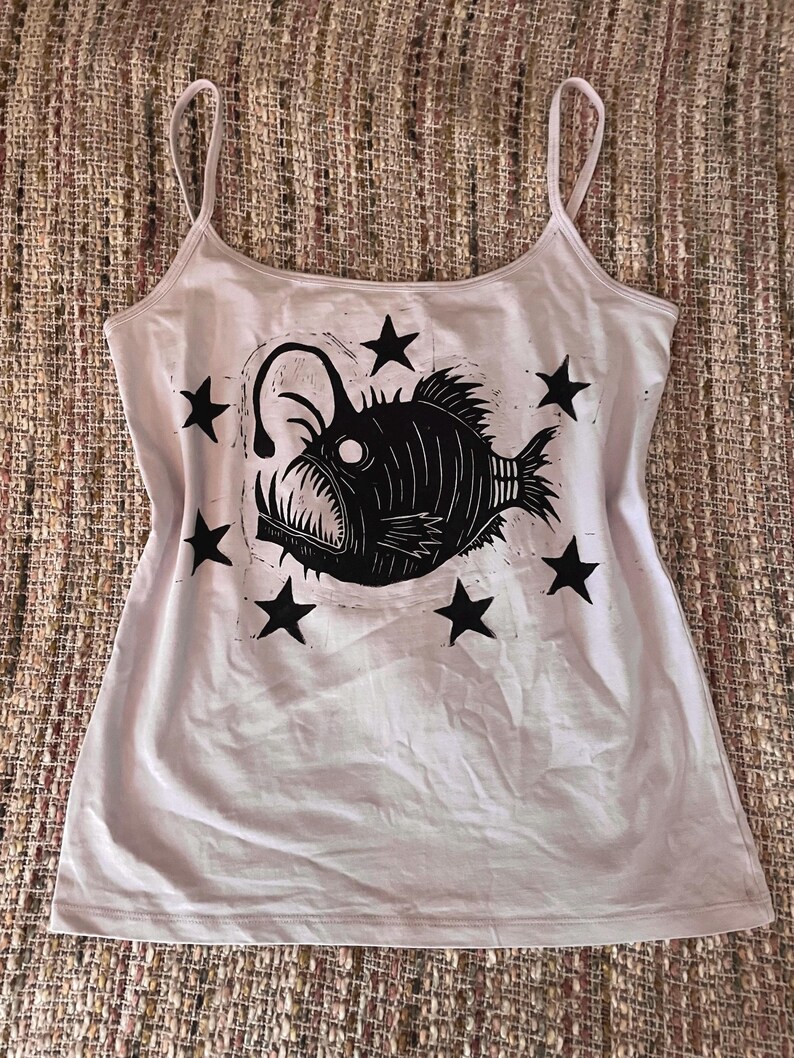 May include: A light pink tank top featuring a black graphic print of an anglerfish surrounded by six black stars. The garment has a scoop neckline and thin spaghetti straps, with the fish illustration centered on the front of the shirt.