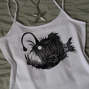 May include: A white tank top featuring a black graphic print of an anglerfish with a prominent lure and sharp teeth. A silver-toned starfish pendant hangs from a black cord, positioned above the fish graphic on the white fabric of the top.