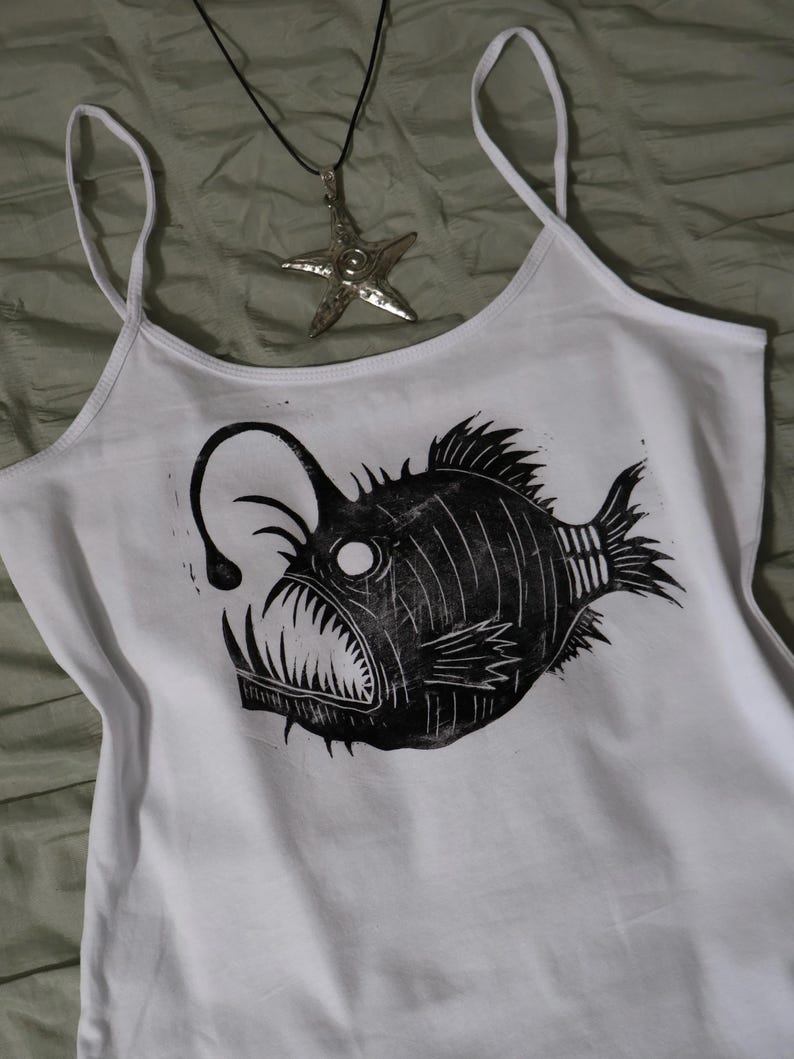 May include: A white tank top featuring a black graphic print of an anglerfish with a prominent lure and sharp teeth. A silver-toned starfish pendant hangs from a black cord, positioned above the fish graphic on the white fabric of the top.