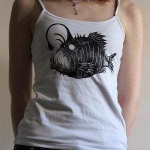 May include: A white spaghetti strap tank top featuring a black graphic print of an anglerfish. The fish illustration displays a prominent lure, sharp teeth, and textured fins, centered on the front of the garment with a scoop neckline.