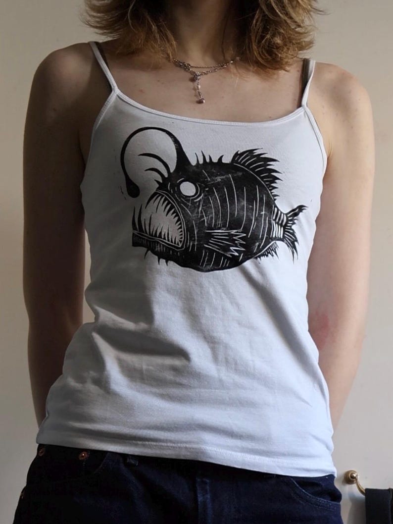 May include: A white spaghetti strap tank top featuring a black graphic print of an anglerfish. The fish illustration displays a prominent lure, sharp teeth, and textured fins, centered on the front of the garment with a scoop neckline.