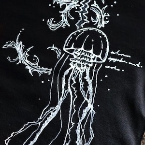 May include: A black t-shirt featuring a white line-art illustration of a jellyfish with long, flowing tentacles. The design includes decorative swirling patterns and illegible cursive script text surrounding the central marine creature.