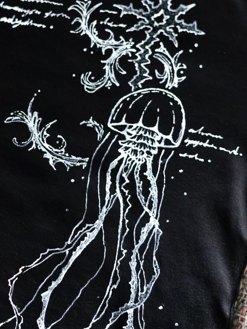 May include: A black t-shirt featuring a white line-art illustration of a jellyfish with long, flowing tentacles. The design includes decorative swirling patterns and illegible cursive script text surrounding the central marine creature.