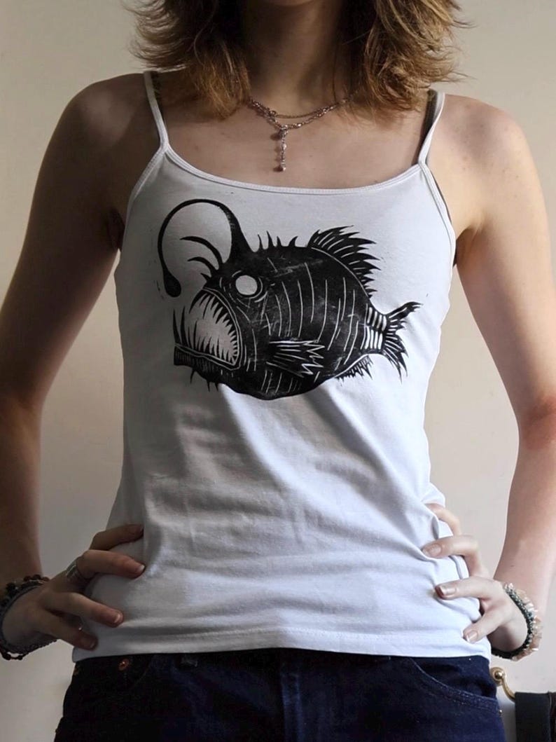 May include: A white spaghetti strap tank top featuring a black graphic print of an anglerfish. The fish illustration displays a prominent lure, sharp teeth, and textured fins, centered on the front of the garment. The top has a scoop neckline.