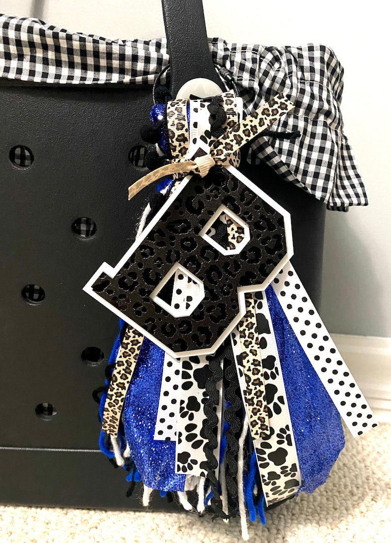 Puede incluir: Un abalorio con la letra "B" en blanco y negro con un dise&ntilde;o de estampado de leopardo. El abalorio est&aacute; unido a un borla azul, blanco y negro con cinta de estampado de leopardo y cinta de estampado de pata.