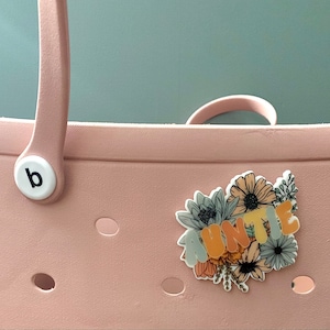 May include: A light pink tote bag with a white 'b' logo and a floral sticker that says 'AUNTIE'. The sticker features a bouquet of flowers in shades of orange, yellow, and blue. The bag has a handle and several circular cutouts.
