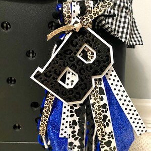 Puede incluir: Un abalorio con la letra B en blanco y negro con un dise&ntilde;o de estampado de leopardo. El abalorio est&aacute; unido a un borla azul, blanco, negro y marr&oacute;n con una variedad de patrones que incluyen huellas de patas y lunares.