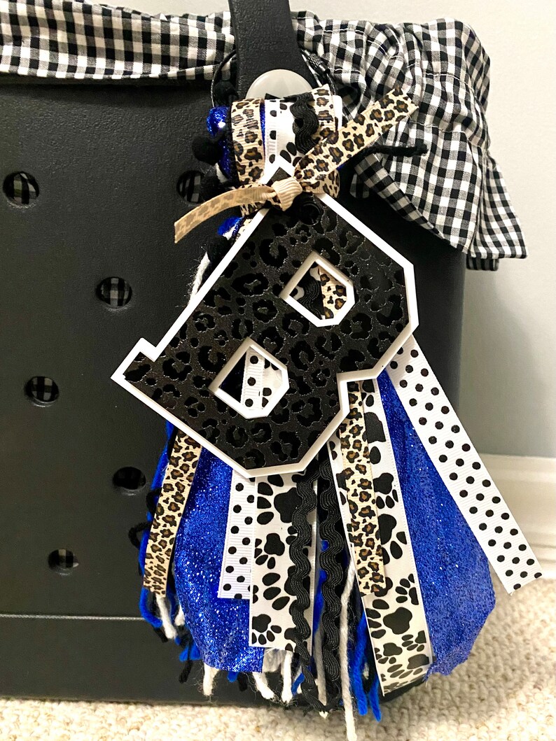 Puede incluir: Un abalorio con la letra B en blanco y negro con un dise&ntilde;o de estampado de leopardo. El abalorio est&aacute; unido a un borla azul, blanco, negro y marr&oacute;n con una variedad de patrones que incluyen huellas de patas y lunares.