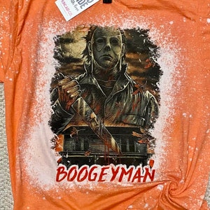 Könnte beinhalten: Orangefarbenes T-Shirt mit einem Design aus Bleichmittelspritzern, das ein Grafikbild von Michael Myers aus der Halloween-Franchise zeigt, der ein Messer hält. Der Text "Boogeyman" ist unter dem Grafikbild gedruckt. Das Shirt hat ein Etikett mit der Aufschrift "Hand Made with love" und "KINGCAD CO."