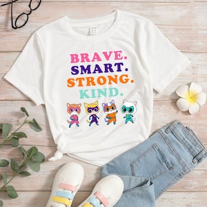 May include: White t-shirt with a graphic of four cartoon cats wearing superhero costumes. The text "BRAVE. SMART. STRONG. KIND." is printed on the shirt.